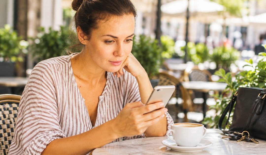A woman with her hair in a bun checks an unbranded small smartphone before meeting a date at a busy but softly blurred outdoor café.
